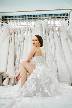 Eine junge brünette Braut sitzt auf einem Bett und starrt auf ein Regal mit Brautkleidern in einem Brautsalon.