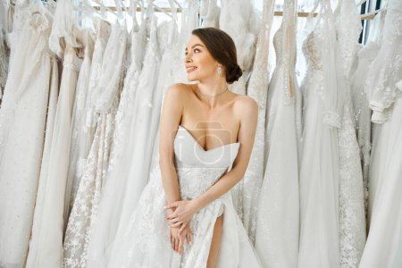 Eine junge, schöne Braut steht in einem Brautsalon vor einem Regal mit weißen Brautkleidern und wählt ihr Kleid sorgfältig aus.
