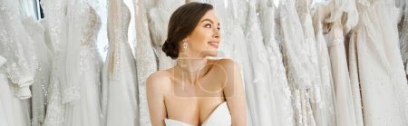 Une jeune, belle mariée brune se tient au milieu d'un rack de robes de mariée blanches dans un salon de mariée.