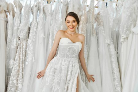 A young brunette bride stands surrounded by a rack of dresses in a wedding salon, looking for her perfect gown.