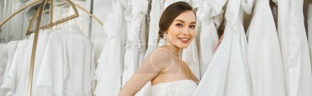 Eine junge, schöne zukünftige Braut steht in einem Hochzeitssalon vor einem Kleiderständer und wählt ihr perfektes Kleid.