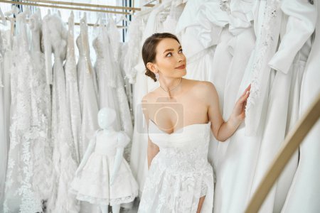 Eine junge, schöne Brünette steht in einem Hochzeitssalon vor einem Kleiderständer und wählt ihr perfektes Kleid.