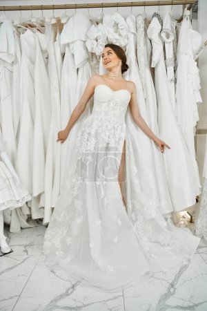 A young, beautiful bride admires a rack of dresses in a wedding salon, contemplating her perfect gown.