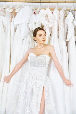 A young brunette bride stands in a wedding salon, surrounded by a rack of beautiful dresses, contemplating her options.