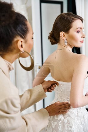 A young brunette bride in a white dress helping a woman put on earrings in a wedding salon.