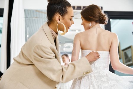 Foto de Una joven novia morena en un vestido blanco y una dama de honor en un traje beige se unen en un salón de bodas, exudando elegancia. - Imagen libre de derechos