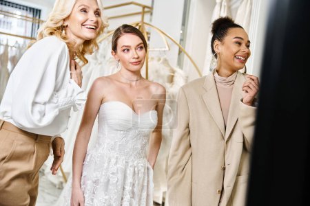 Drei Frauen, eine junge Brünette und eine atemberaubende Braut in einem weißen Kleid stehen gemeinsam vor einem Spiegel.