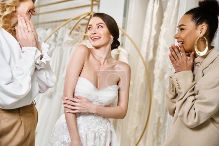 Eine junge brünette Braut in einem weißen Kleid steht neben zwei anderen Frauen und strahlt Eleganz und Anmut aus.