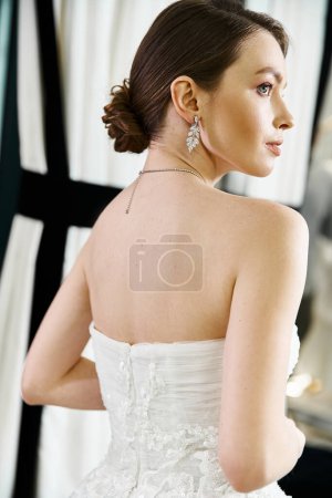 A young brunette bride in a white wedding dress admiring her reflection in a mirror at a wedding salon.