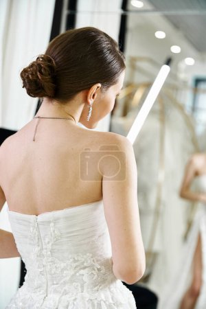 A young brunette bride in a white wedding dress gazes at her reflection in a mirror in a bridal salon.