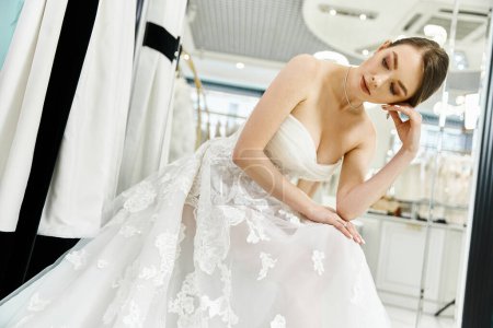 A young, beautiful brunette bride in a stunning white wedding dress gazes at her reflection in a mirror.