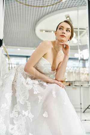 A young brunette bride in a flowing white dress sits elegantly on a chair in a wedding salon.