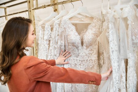 Una joven novia hermosa está examinando cuidadosamente un estante de vestidos de novia en una boutique nupcial.