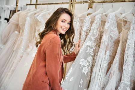 Una joven novia hermosa de pie delante de un estante de vestidos de novia elegantes, cuidadosamente seleccionando el perfecto.