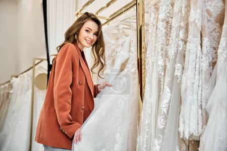 Una joven novia hermosa está de pie delante de un estante de vestidos de novia, examinando cuidadosamente sus opciones