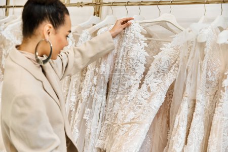 Eine junge, schöne Braut wählt mit Hilfe einer Verkäuferin sorgfältig Brautkleider aus einem vielfältigen Regal aus.