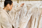 A young, beautiful bride carefully selecting wedding dresses from a diverse rack with the assistance of a shop attendant. Sweatshirt #698530302