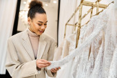 A young beautiful bride gazes at a wedding dress on a rack, smiling as she looking at one Poster #698530358