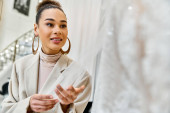 A young bride shops for her wedding dress, standing in front of a mirror and gowns mug #698530360