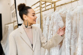 A young beautiful bride is carefully inspecting a rack filled with elegant wedding dresses Poster #698530368