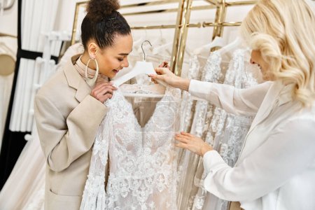 Two young women, a beautiful bride-to-be and a shop assistant, delicately examining a dress hanging on a rack in a boutique.