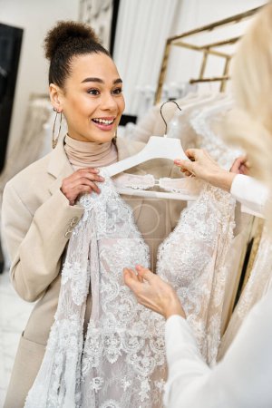Foto de Una joven y hermosa novia explora un vestido en una percha mientras compra para su boda, asistida por un asistente de tienda útil. - Imagen libre de derechos