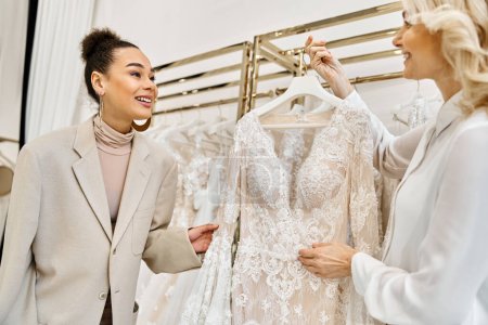 Téléchargez les photos : Jeunes femmes admirant une robe de mariée sur une étagère dans un magasin de mariage, assistées par un commerçant. - en image libre de droit