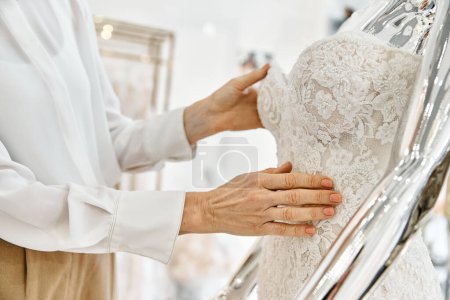 A middle-aged shopping assistant stands confidently in front of a rack of elegant dresses in a wedding salon.