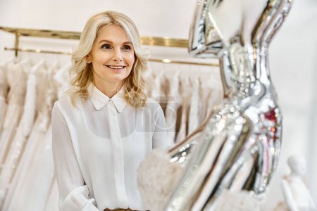 A middle-aged, beautiful shopping assistant stands confidently in front of a rack of elegant dresses in a wedding salon.