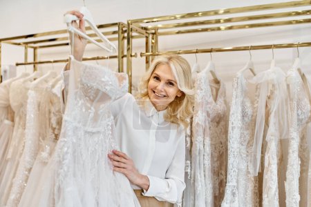 A middle-aged beautiful shopping assistant stands gracefully in front of a rack of elegant wedding dresses in a bridal salon.