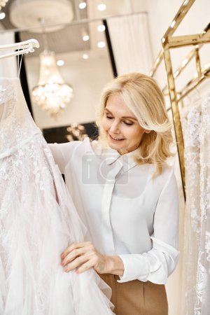 A middle-aged beautiful shopping assistant browses wedding dresses on rack in a bridal salon.