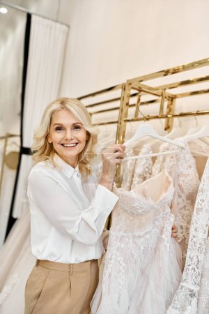 A middle-aged beautiful shopping assistant stands elegantly in front of a rack of wedding dresses in a bridal salon.