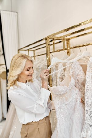 A middle-aged beautiful shopping assistant helps a woman browse through wedding dresses on a rack in a salon.