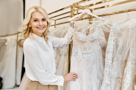 A middle-aged beautiful shopping assistant in a wedding salon stands in front of a rack of dresses, ready to assist customers.