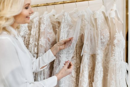 A middle-aged beautiful shopping assistant browses wedding dresses on a rack in a bridal salon.