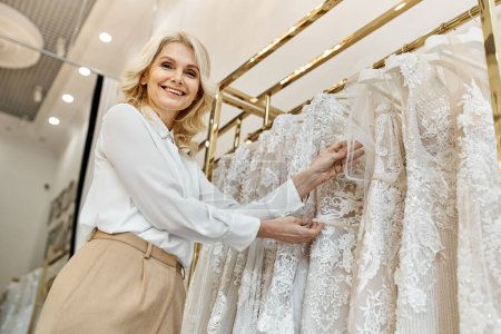 A middle-aged beautiful shopping assistant stands in front of a rack of dresses in a wedding salon, assisting customers.