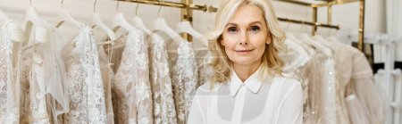 Schöne Verkäuferin mittleren Alters, die vor einem Kleiderständer in einem Hochzeitssalon steht.