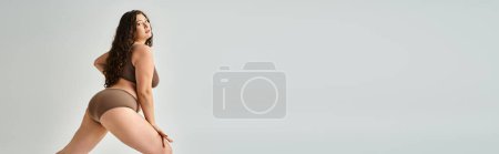 Foto de Estandarte de mujer con curvas en ropa interior con pelo castaño rizado posando de lado sobre fondo gris - Imagen libre de derechos