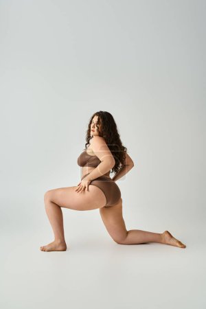 Foto de Atractiva mujer de talla grande con el pelo castaño rizado posando de lado con las piernas sobre fondo gris - Imagen libre de derechos