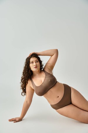 alluring plus size woman in lingerie with curly brown hair leaning on arm and putting hand on head