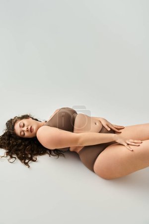 Foto de Seductora mujer joven de talla grande en ropa interior marrón con las manos en las caderas y los ojos cerrados - Imagen libre de derechos