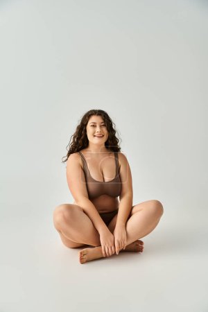 cheerful curvy young woman in brown underwear sitting cross legged against grey background
