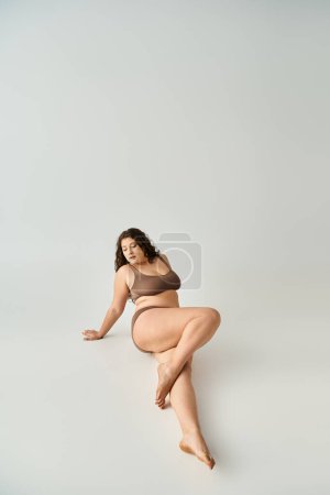 Photo for Alluring plus size young woman in brown underwear sitting, leaning to arms and putting leg - Royalty Free Image
