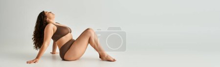 Téléchargez les photos : Bannière de séduisante fille courbée en sous-vêtements marron assise, penchée vers les bras et tirant les jambes vers elle - en image libre de droit