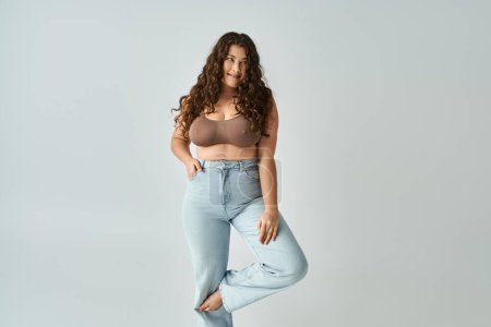 Photo for Smiling plus size young woman in brown bra and blue jeans with curly hair posing with bent leg - Royalty Free Image