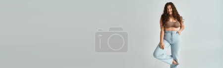 Photo pour Bannière de femme gaie plus la taille en soutien-gorge marron et jeans bleus aux cheveux bouclés posant avec jambe courbée - image libre de droit