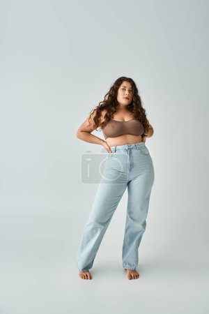 lovely curvy girl in brown bra and blue jeans posing with hands in pockets on grey background