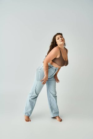 beautiful plus size woman in brown bra and blue jeans leaning to forward on grey background