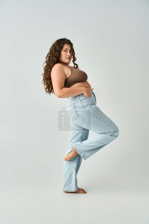 pretty curvy woman in brown bra and blue jeans leaning to behind with bent leg on grey background