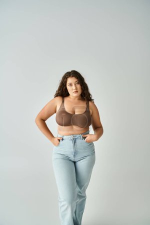 cheerful plus size woman in brown bra and blue jeans posing with hands in pockets on grey background